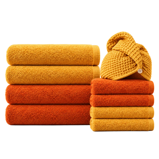 A set of folded towels in warm autumn shades, including mustard yellow, orange, and rust red, with a matching waffle-texture turban hat on top, arranged on a light gray background.